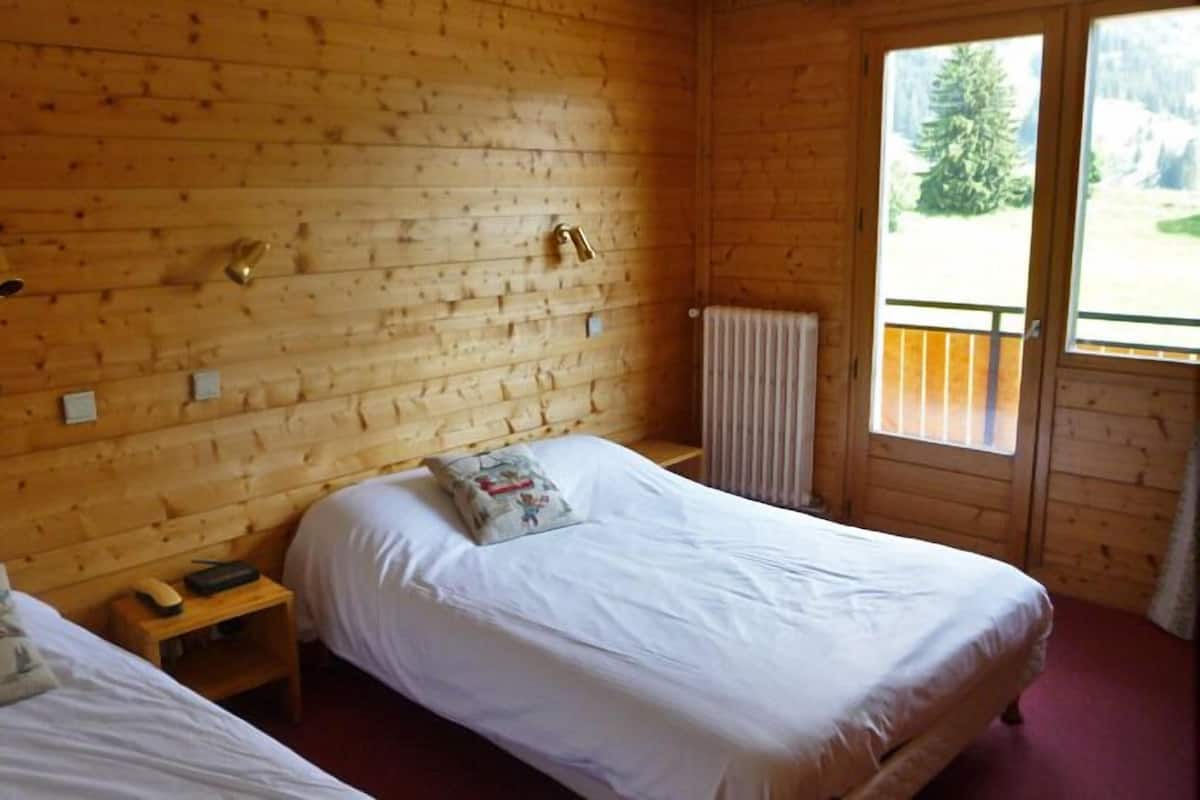 Una habitación de cabaña de madera con dos camas, una mesita y un ventanal con vistas a árboles.