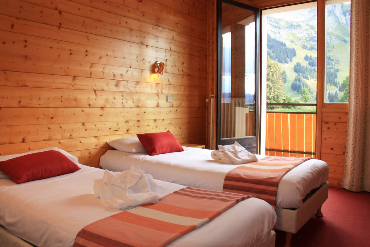 Habitación de cabaña de madera con dos camas, un almohadón rojo y vista a un paisaje montañoso a través de un ventanal.