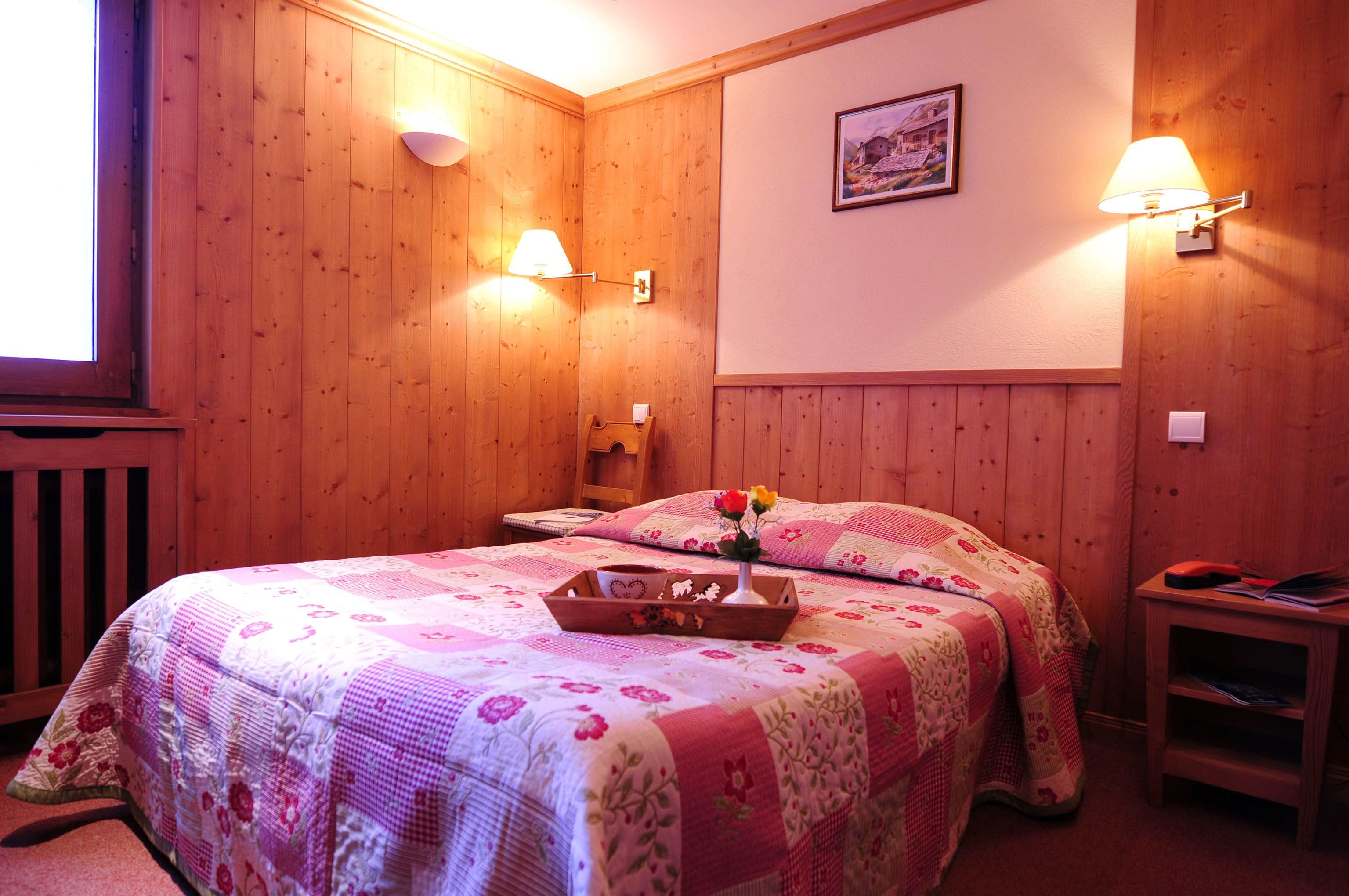 Una habitación con paneles de madera, una cama, dos lámparas fijadas a la pared, una mesita pequeña y un cuadro enmarcado.