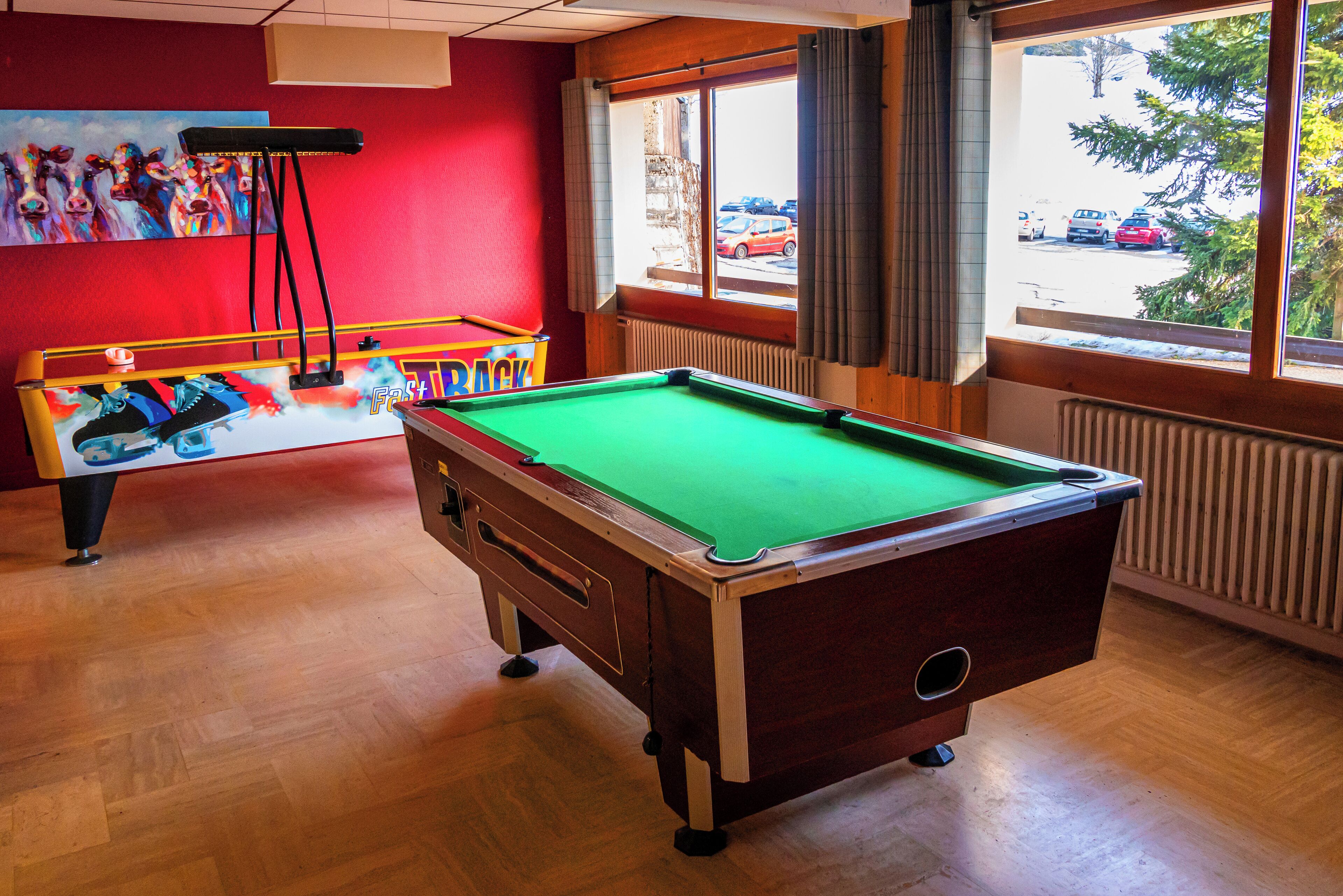 Une table de billard dans une pièce au plancher en bois, avec un mur rouge et une fenêtre donnant sur les voitures garées à l’extérieur.