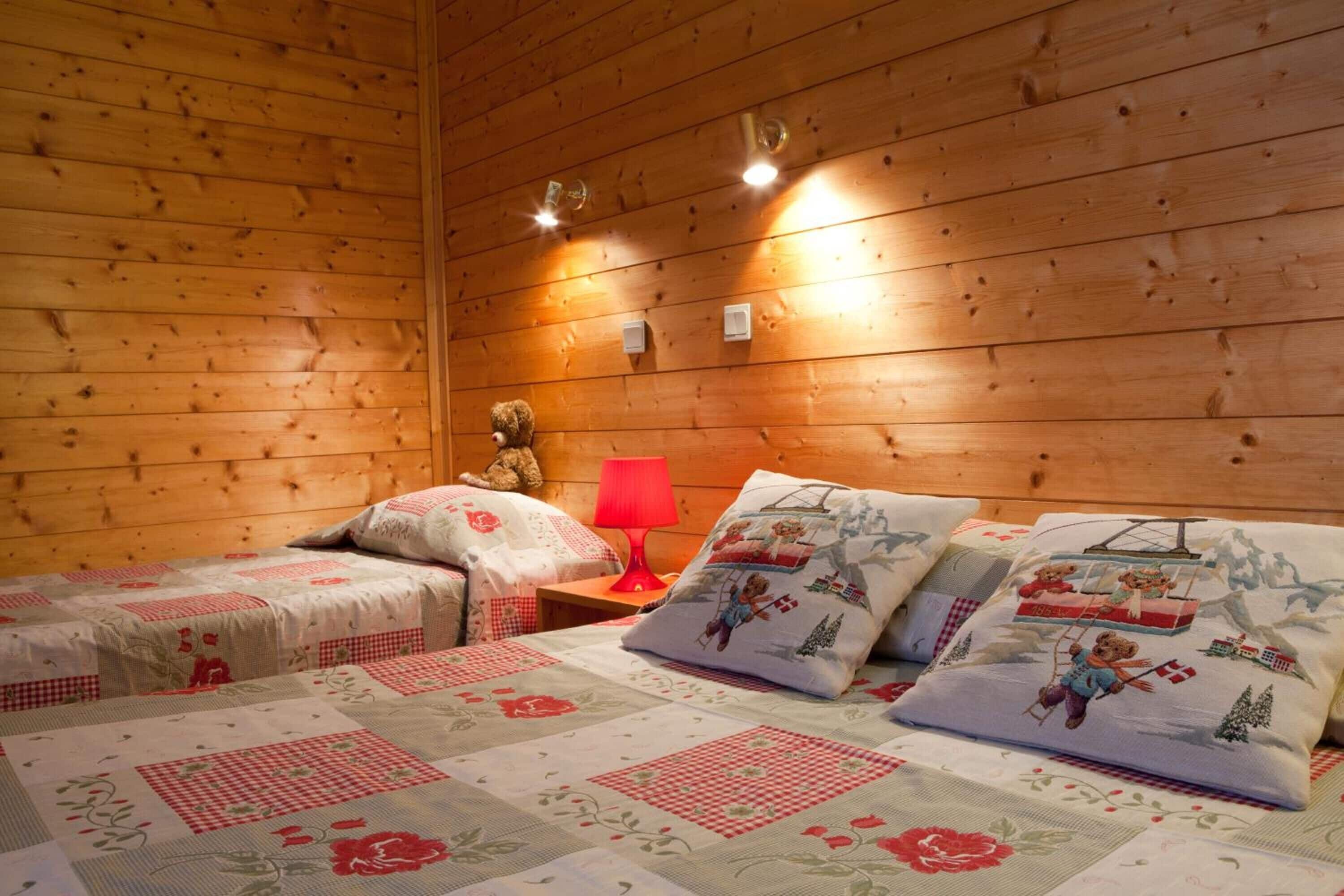 Une pièce chaleureuse aux murs en bois, avec deux lits dotés de draps à motifs floraux, une lampe rouge et des ours en peluche.