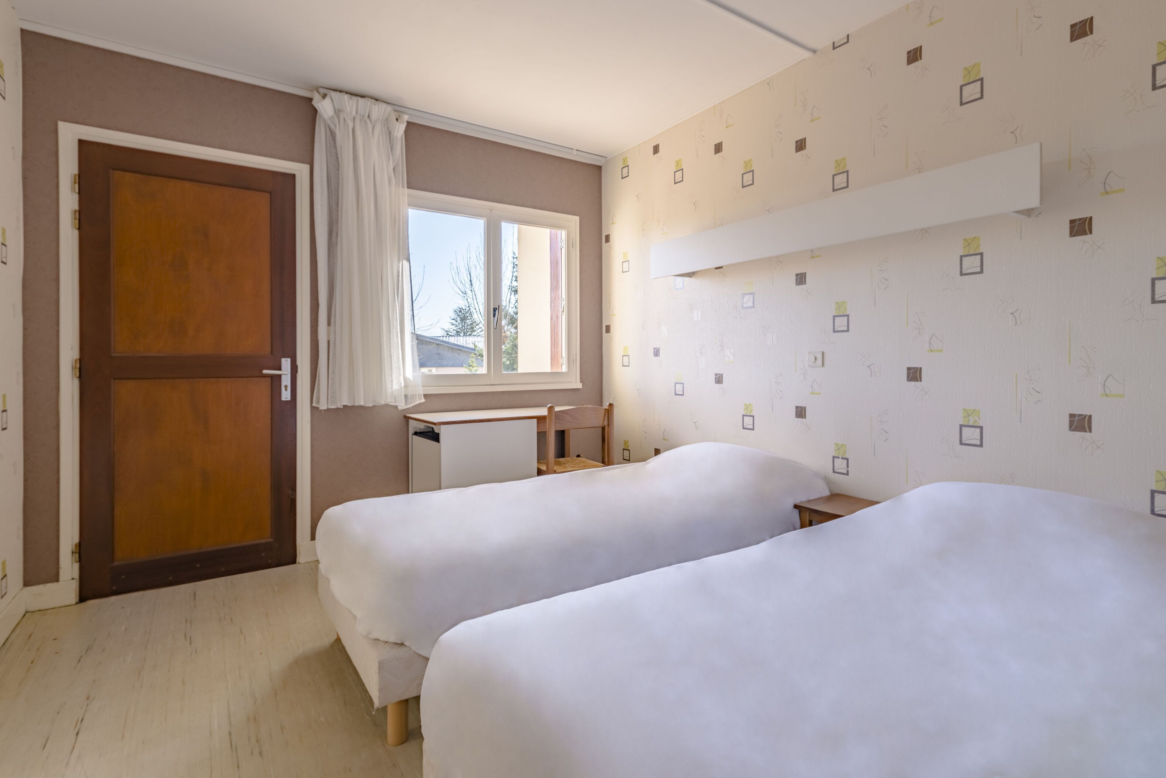 A room with two beds, a wooden door, a window with curtains, a desk, and a patterned wall.
