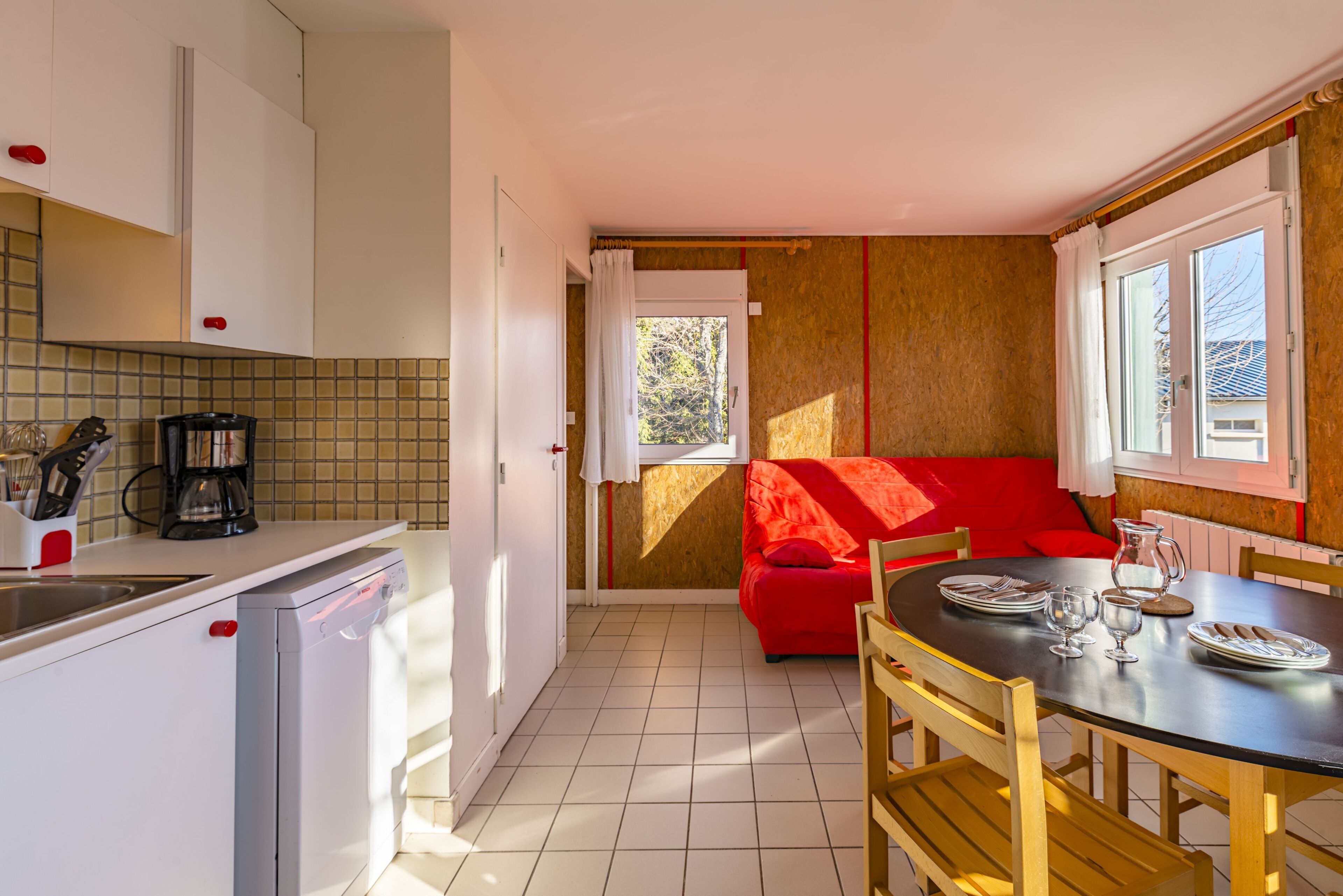 A compact kitchen with a dining area, a red sofa, and a window with curtains.