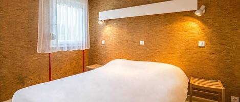 A bedroom with a large bed, a bedside table, a chair, a window with sheer curtains, and wall-mounted lighting.