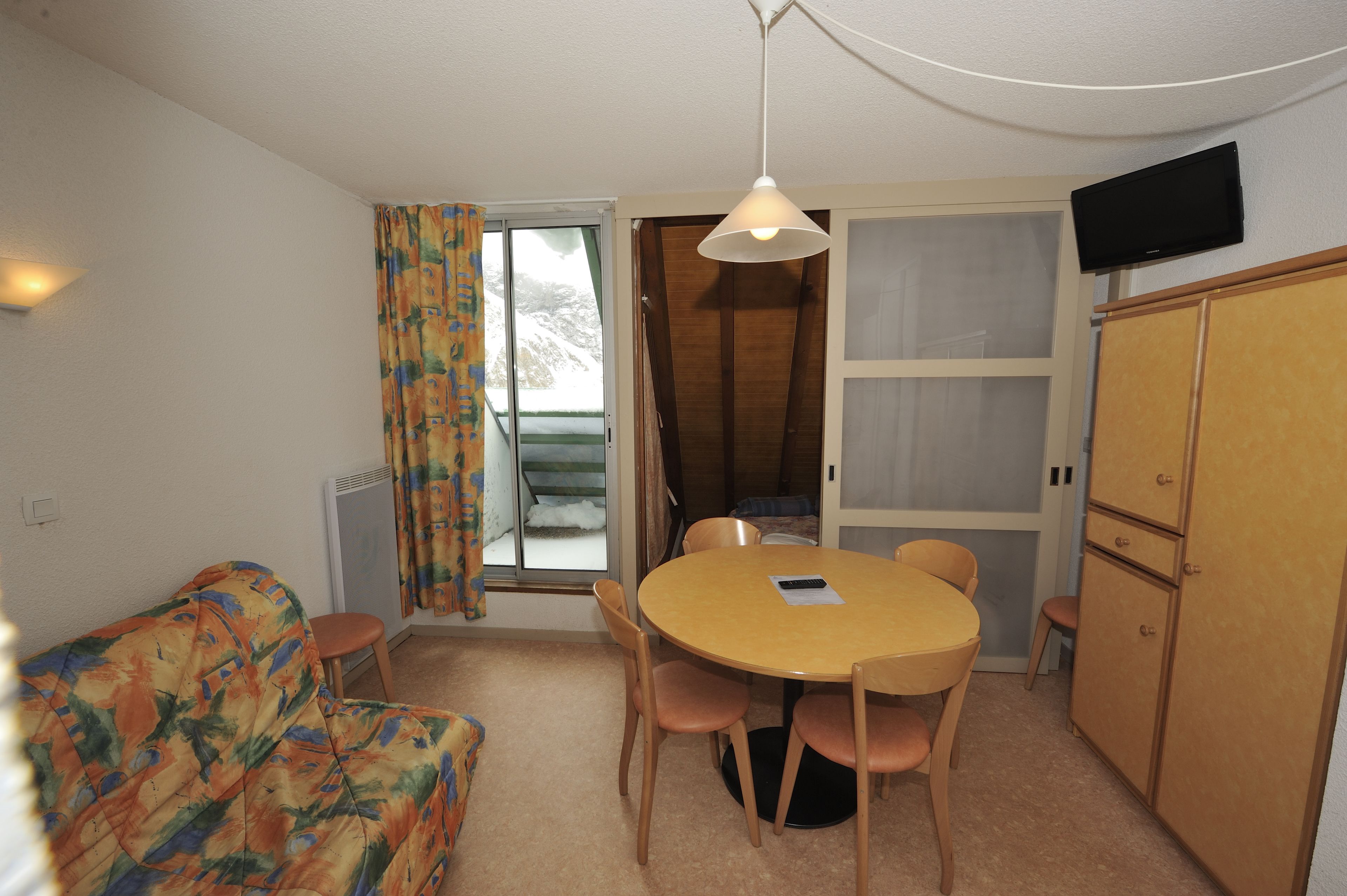 Ein kompaktes Zimmer mit einer Couch, einem runden Tisch mit Stühlen, einem an der Wand befestigten Fernseher und einer Schiebetür, die auf einen Balkon führt.