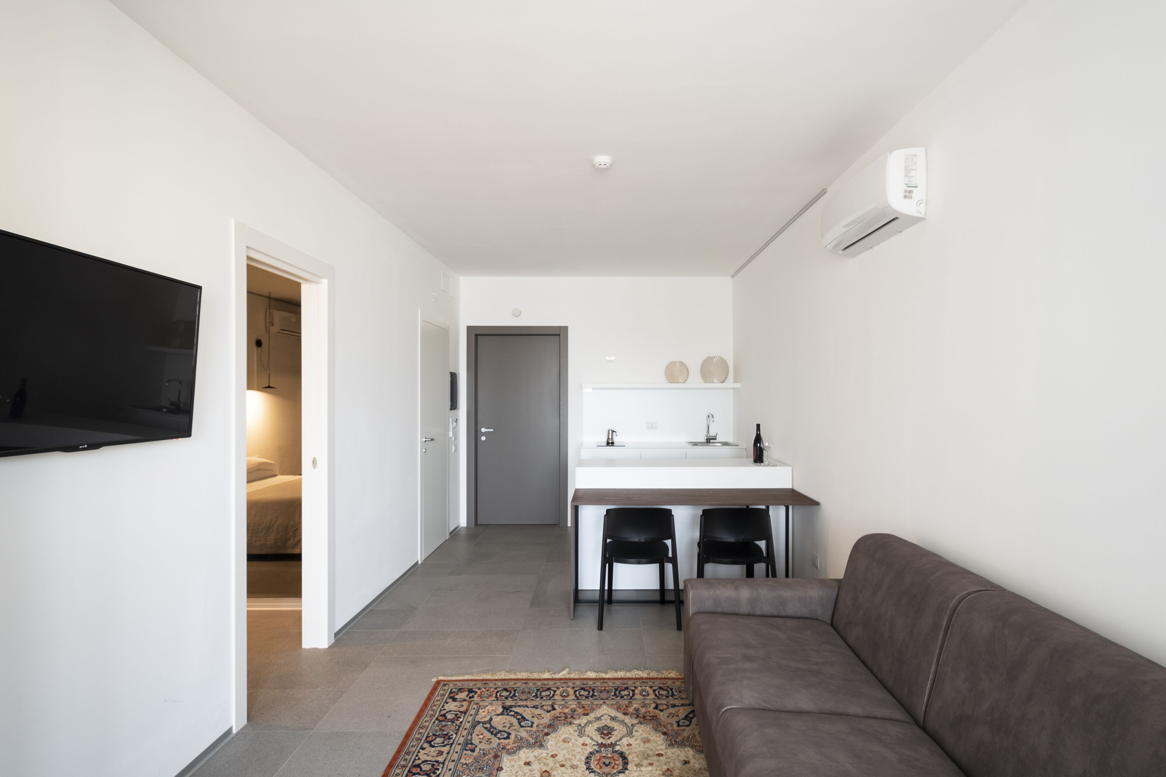 Uma sala de estar moderna com um sofá cinzento, uma televisão de tela plana na parede e uma área de cozinha com bancada branca e duas cadeiras pretas.