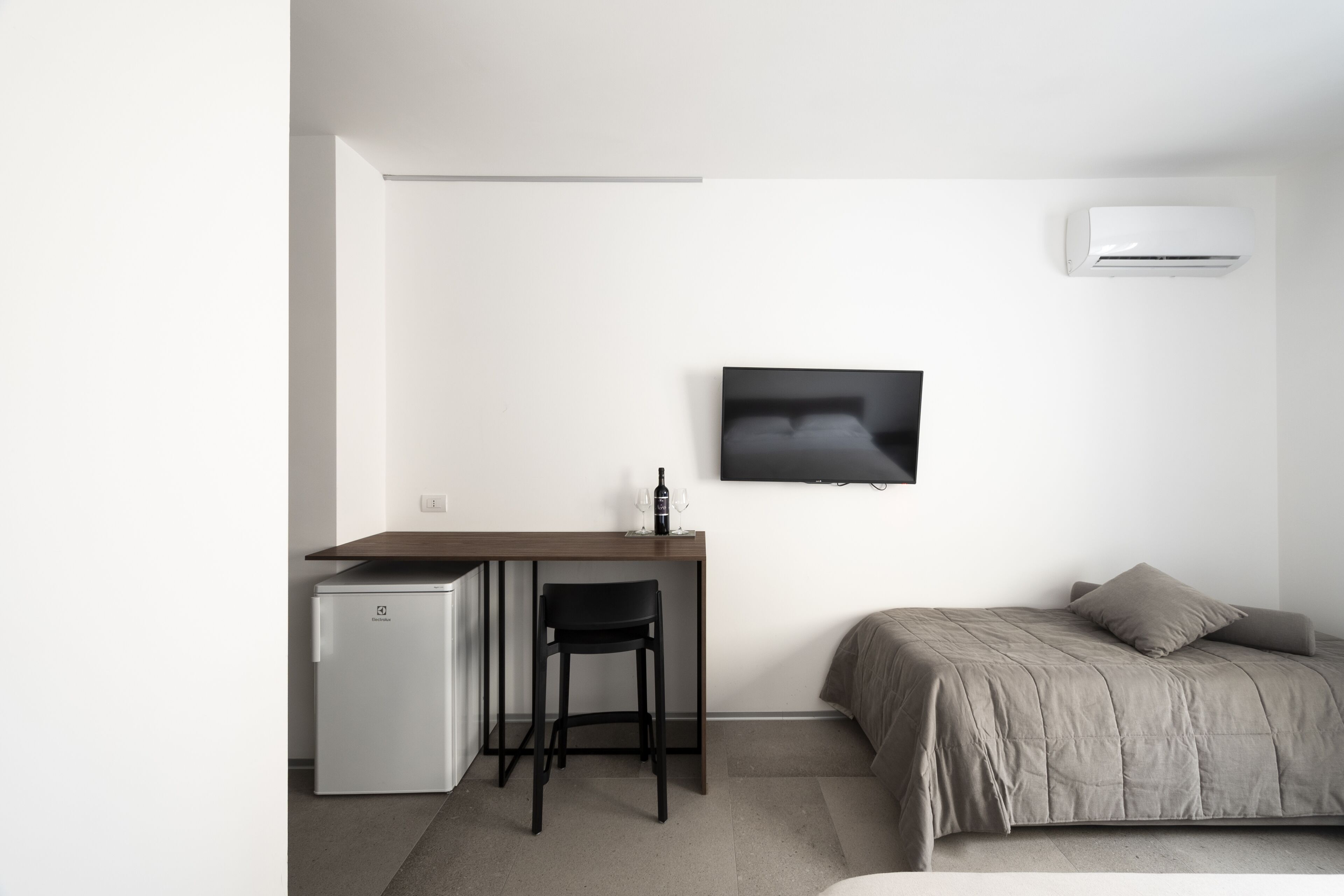 Un dormitorio minimalista con una cama, un escritorio pequeño con una silla, un televisor y un refrigerador compacto.