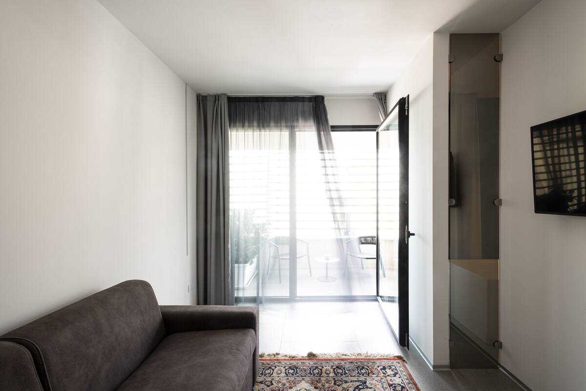 A modern living room with a sofa, a patterned rug, and a sliding glass door leading to a balcony with outdoor furniture.