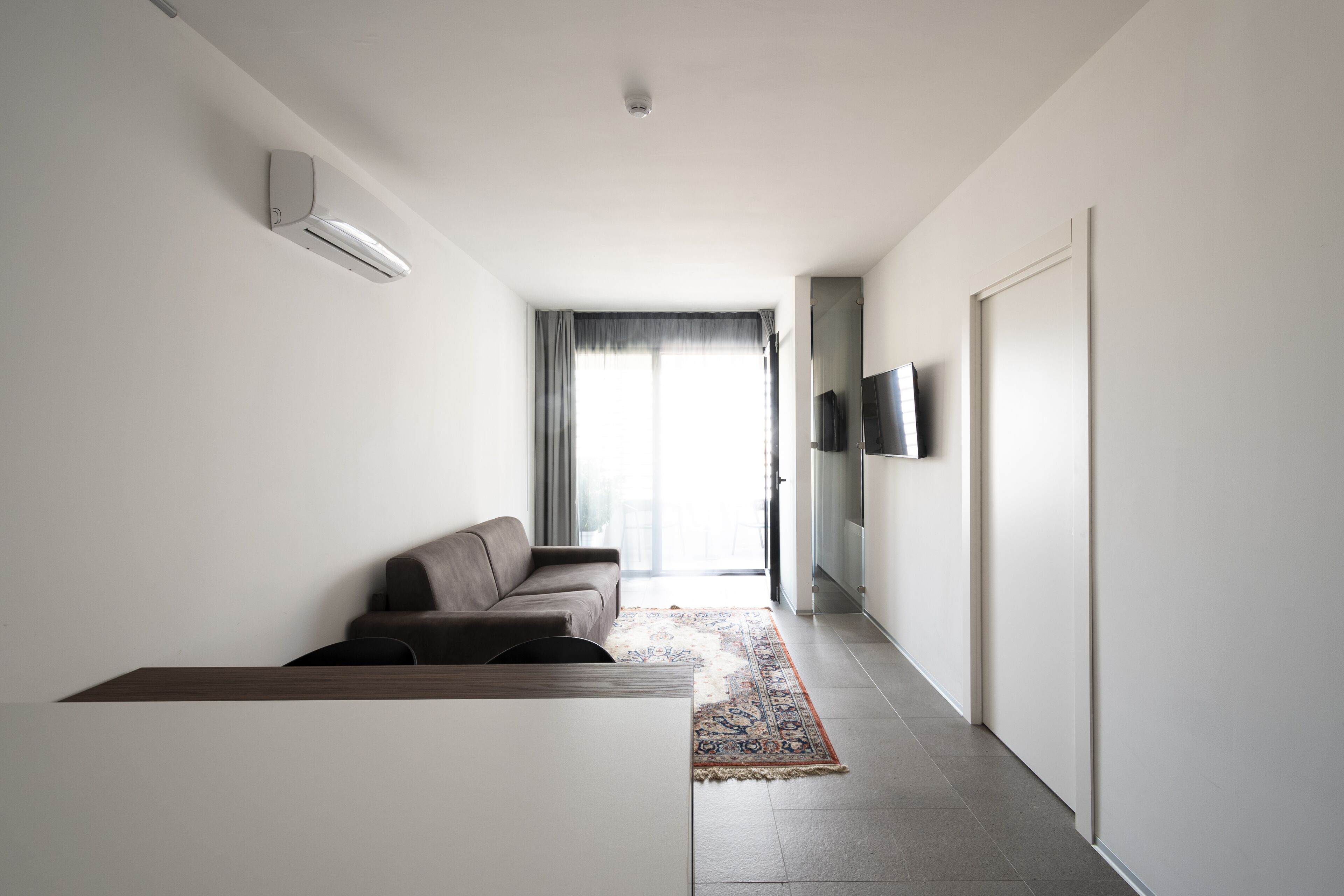 A modern living room with a sofa, a flat-screen TV mounted on the wall, and a large window with curtains.