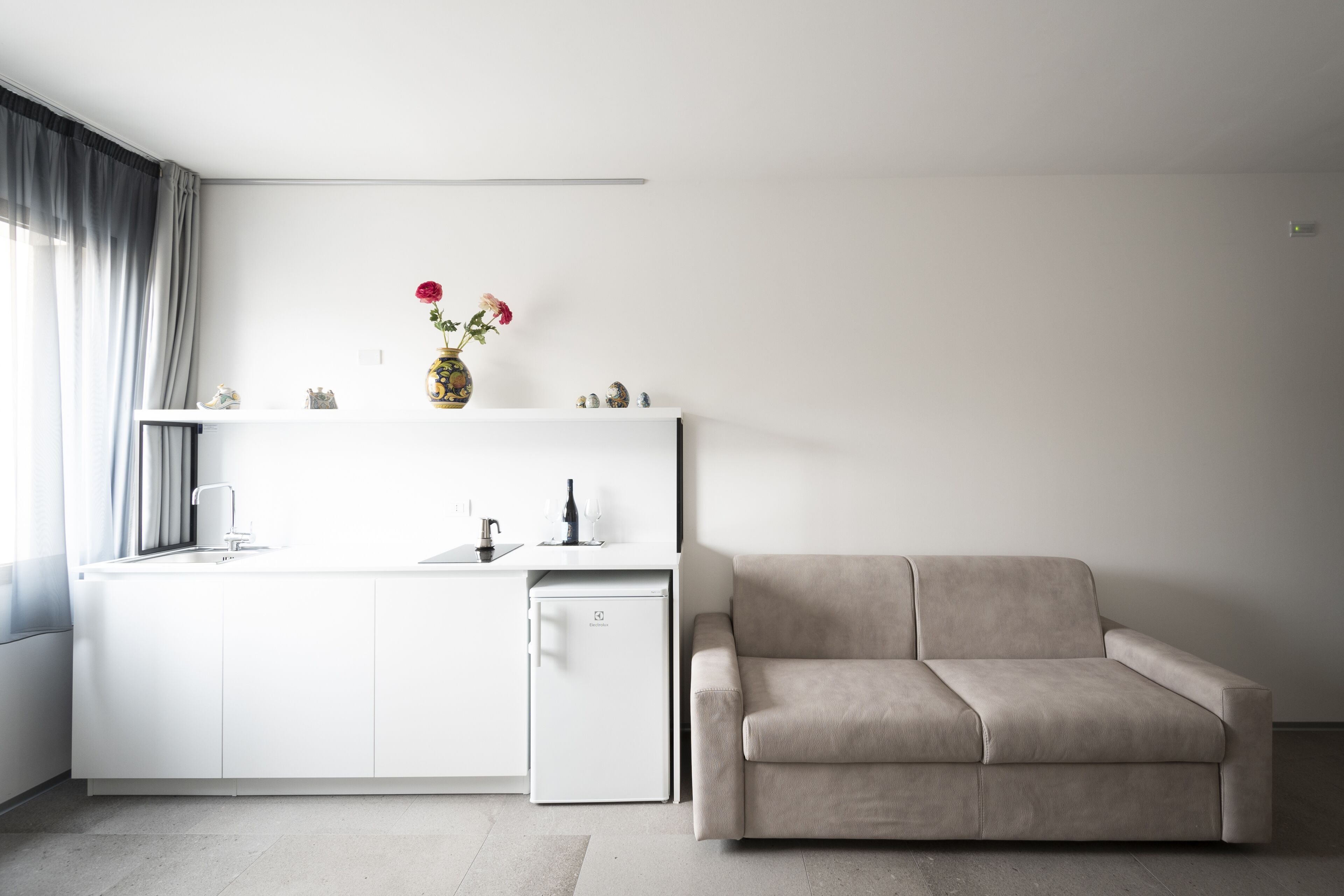 Una cocina moderna con armarios blancos, un refrigerador pequeño y un sofá beige en un espacio habitable minimalista.