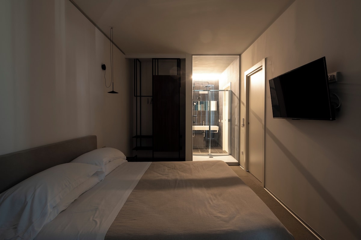 A modern hotel room with a large bed, a TV mounted on the wall, and a shower area visible through an open door.