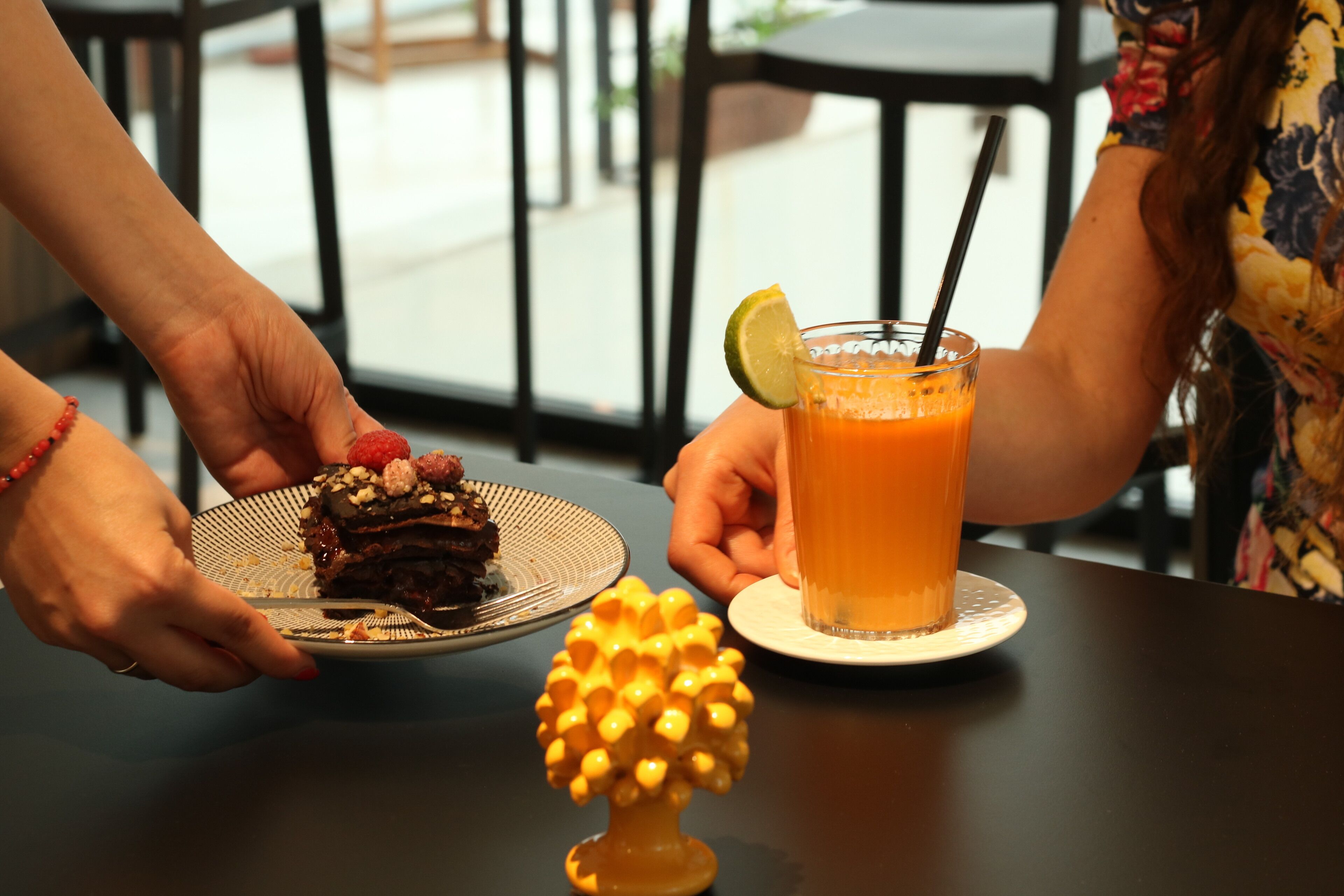 A person holding a plate with a slice of cake and a glass of orange juice with a straw.
