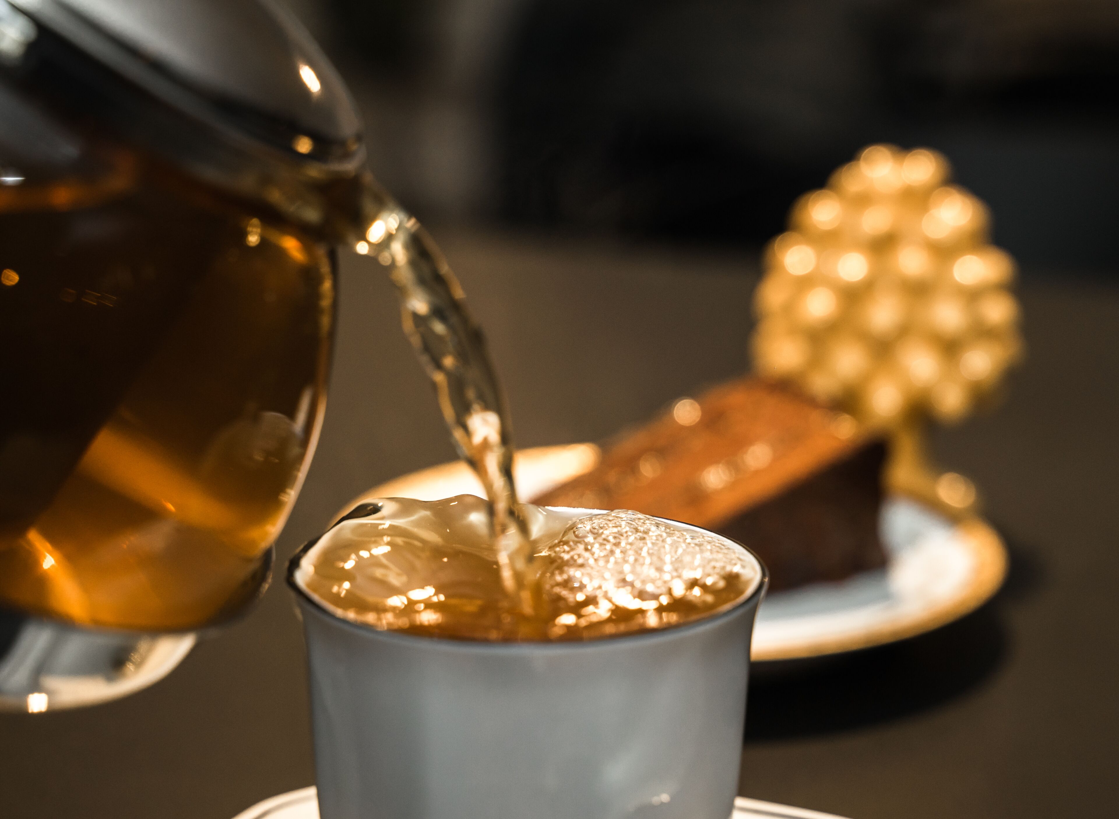 A close-up of a teapot pouring tea into a cup, with a pastry in the background.