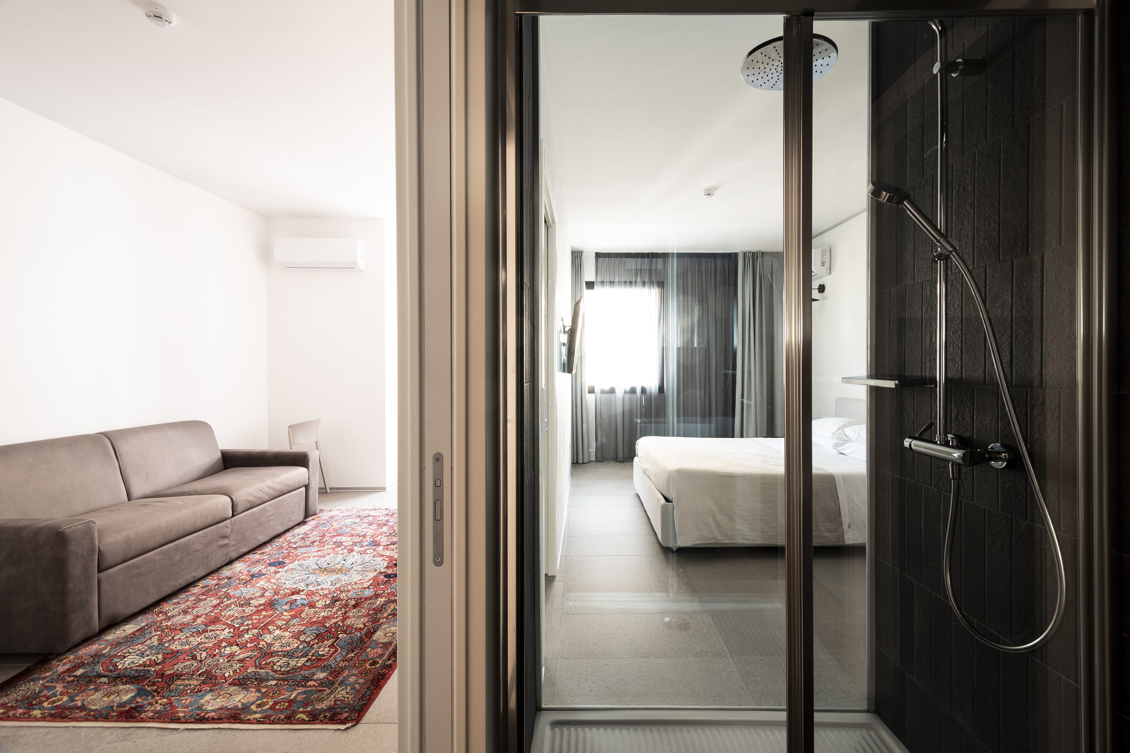 A modern hotel room with a sliding glass door leading to a bathroom with a shower. A sofa and a patterned rug are visible in the living area.