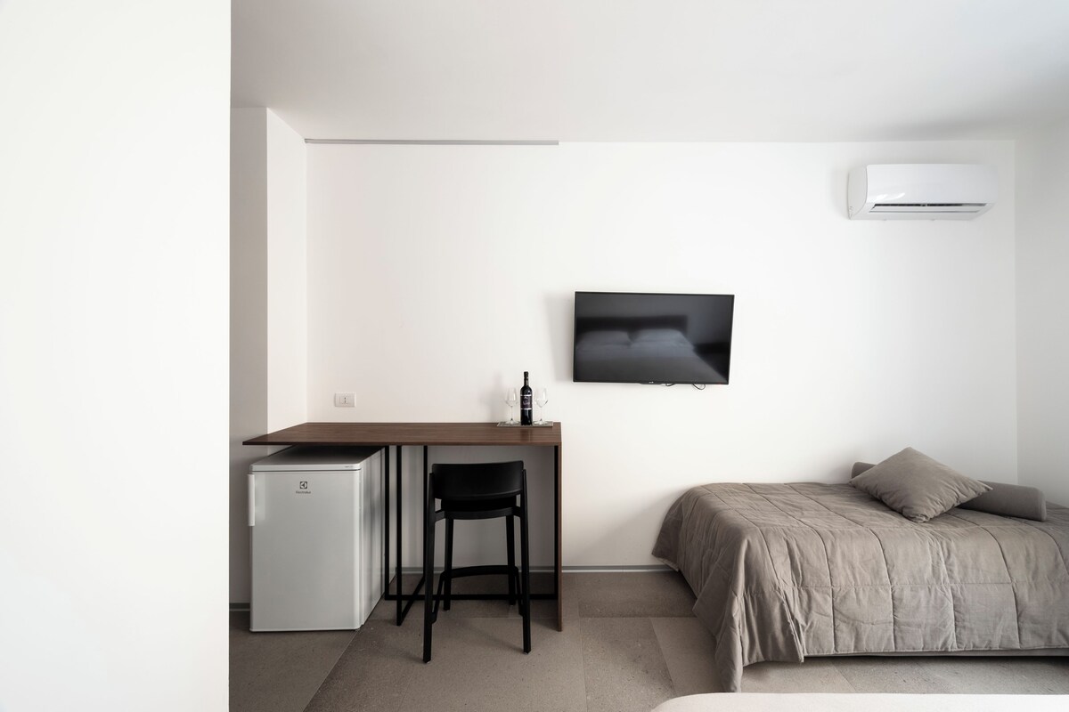 A minimalist bedroom with a bed, a small desk with a chair, a television, and a compact refrigerator.