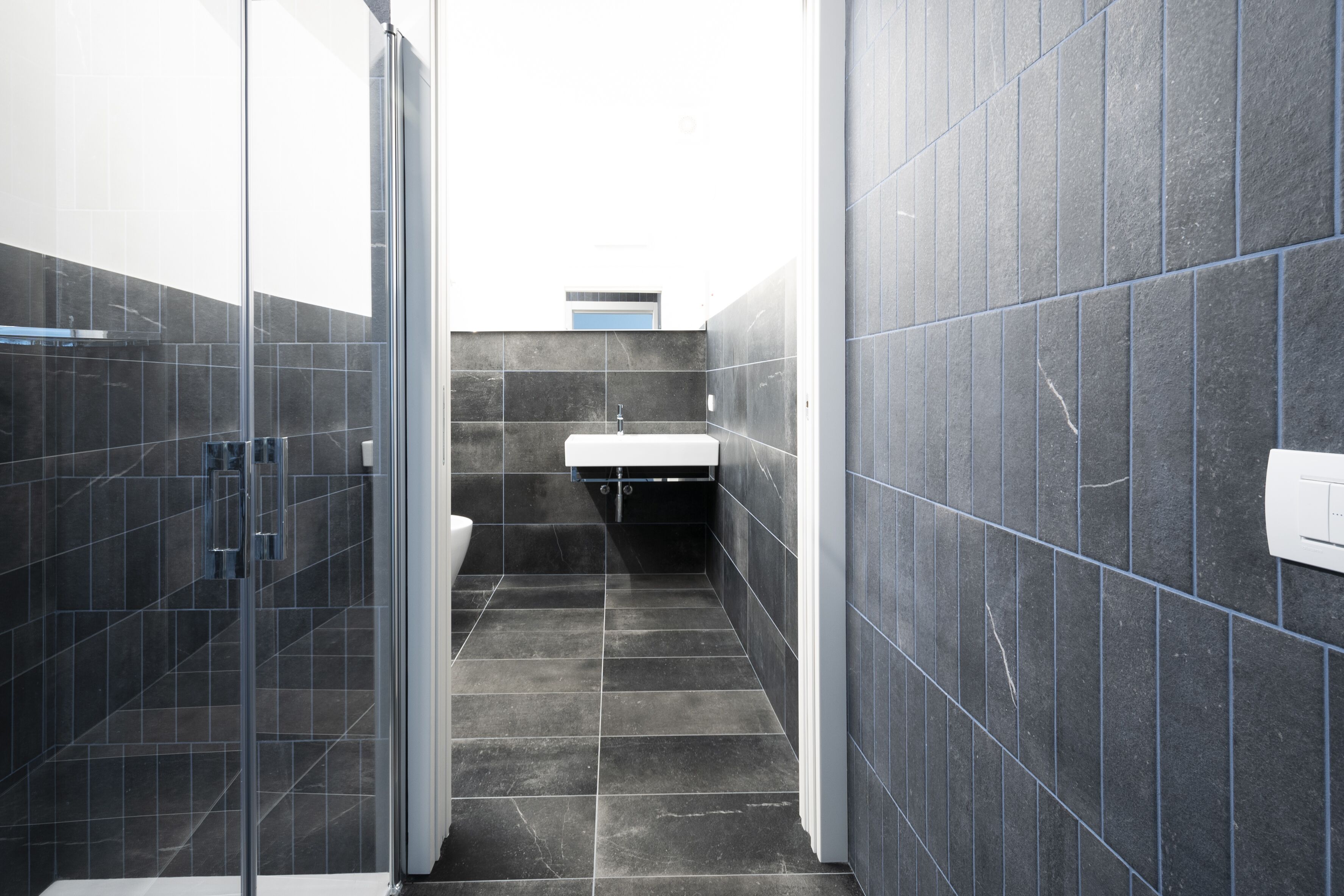 A narrow bathroom with dark tiles, a glass shower enclosure, and a white sink.