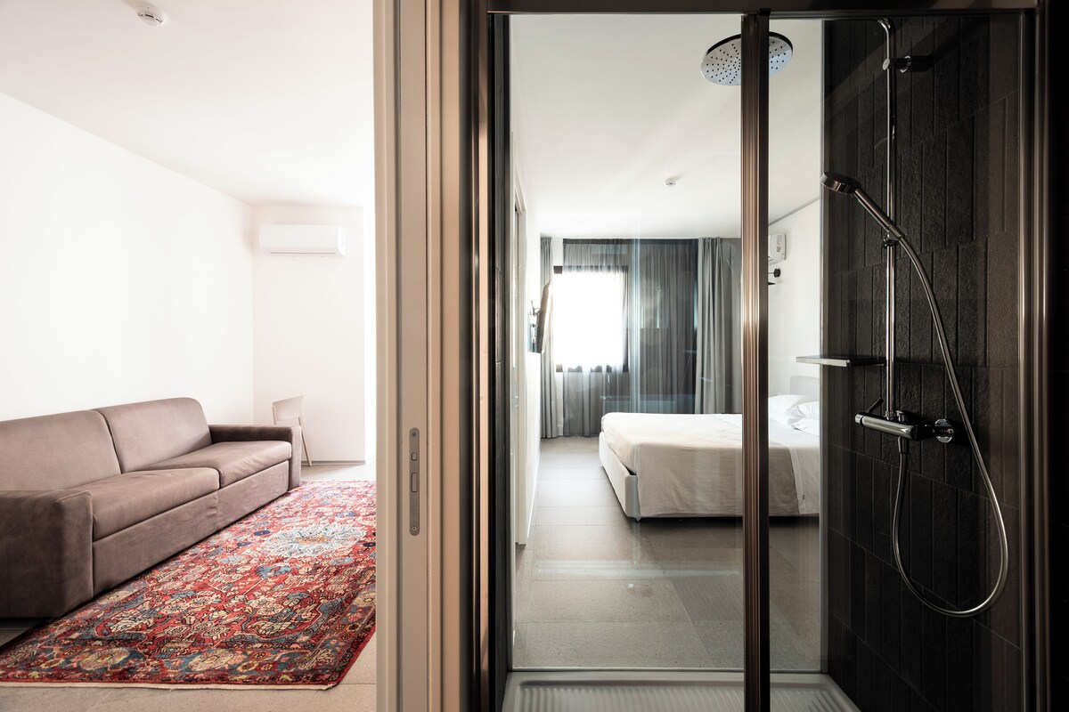 A modern hotel room with a sliding glass door leading to a bathroom with a shower. A sofa and a patterned rug are visible in the living area.