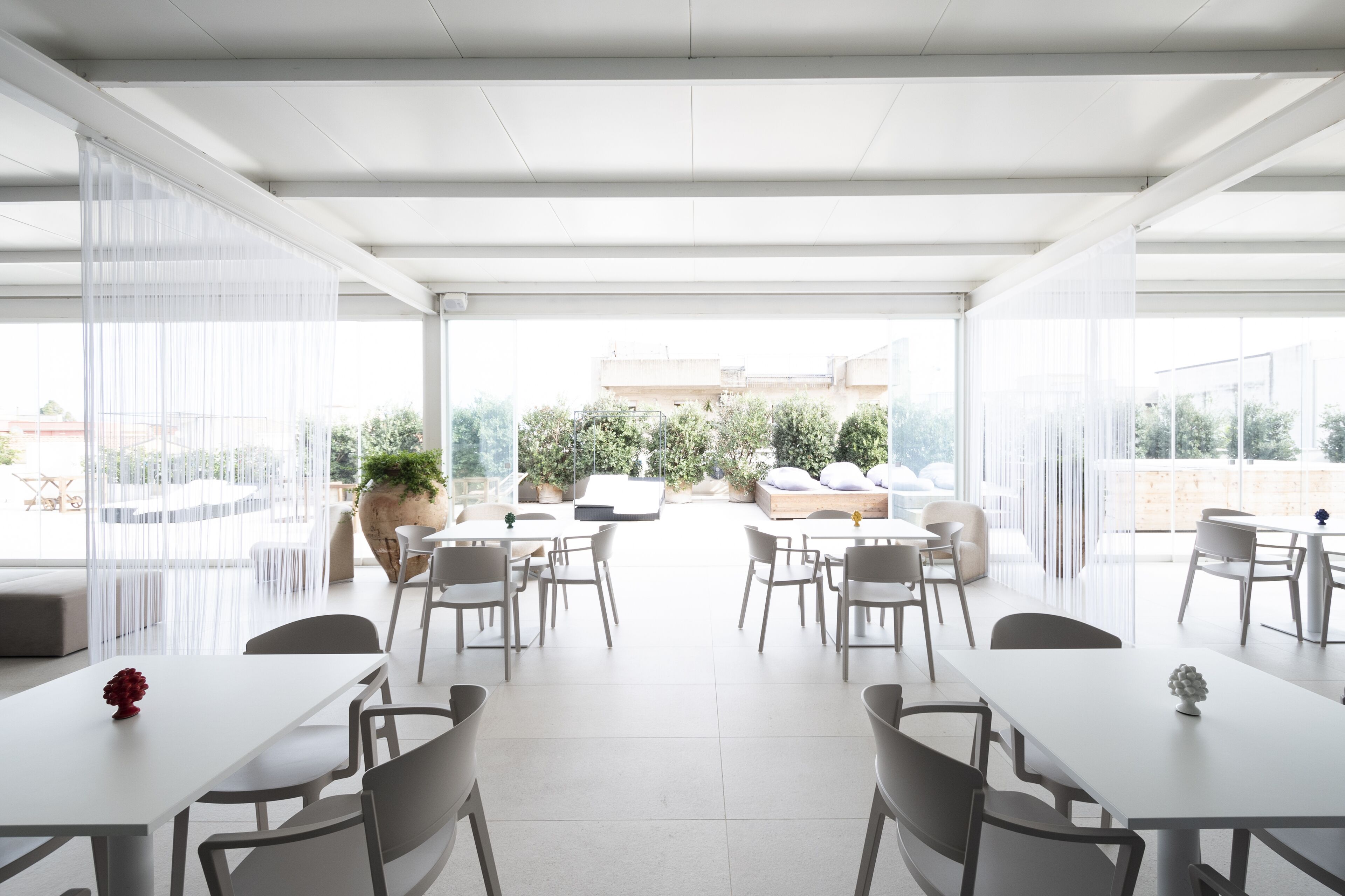 Un espace de restauration moderne, à aire ouverte, avec des tables et des chaises blanches, de grandes fenêtres et une vue sur la verdure et les bâtiments en arrière-plan.