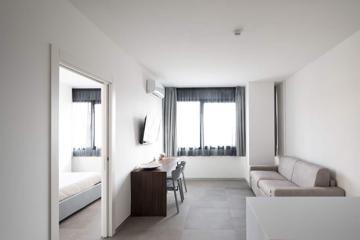 A modern, minimalist room with a bed, a desk with chairs, a sofa, and large windows with curtains.