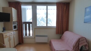 Une petite pièce, simplement aménagée, avec un lit, un bureau, une télévision et une fenêtre avec des rideaux.
