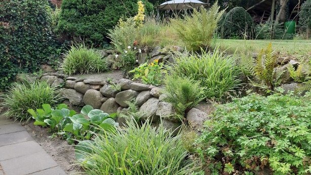 Een tuin met een stenen muur, diverse planten en een prieeltje.