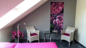 Een intieme kamer met schuine wanden, twee witte rieten stoelen met rode kussens, een klein glazen tafeltje, een groot bloemschilderij en een bed met een roze deken.