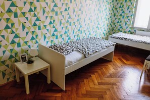 Una habitación con pared de diseño geométrico, suelo de madera en espiga, cama individual con colcha estampada, mesita de noche con lámpara y una planta pequeña, y ventana con persianas.