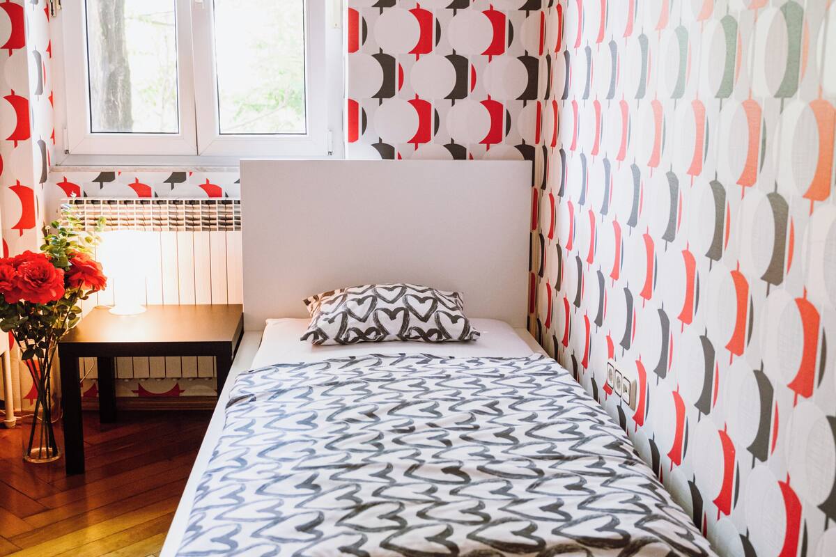 Una habitación con cama, ventana, una mesa con flores y papel tapiz estampado.