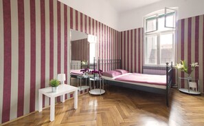 Un dormitorio con papel tapiz a rayas, una cama, una mesita de noche con una lámpara, una silla y una ventana con vistas a edificios.