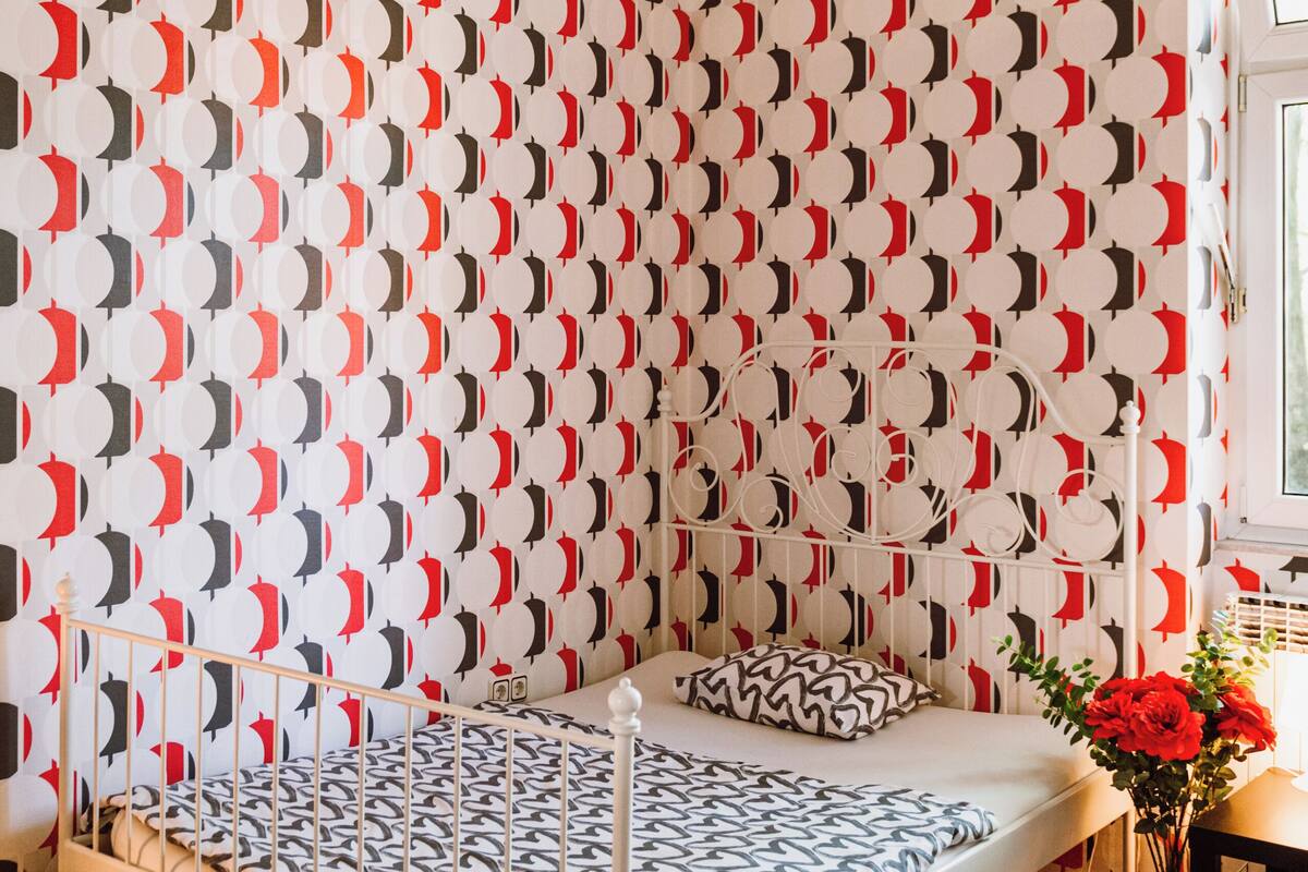 Una habitación con papel tapiz estampado, una cama con estructura de metal, suelo de madera y un ventanal.