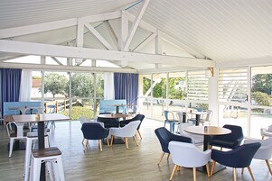 Un vaste espace intérieur doté de nombreuses tables et chaises, de grandes fenêtres offrant une vue sur l’extérieur et d’un plafond haut avec des poutres apparentes.