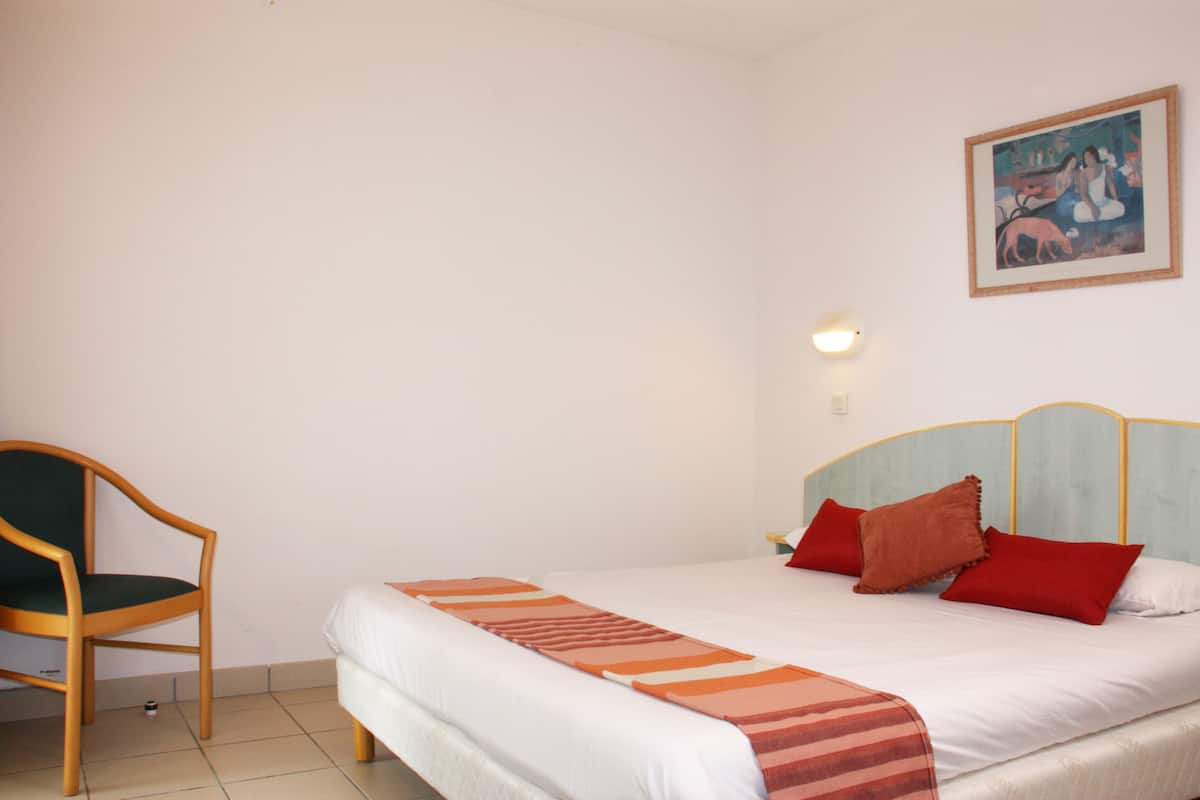 A bedroom with a bed, a chair, a painting on the wall, and a wall-mounted light.