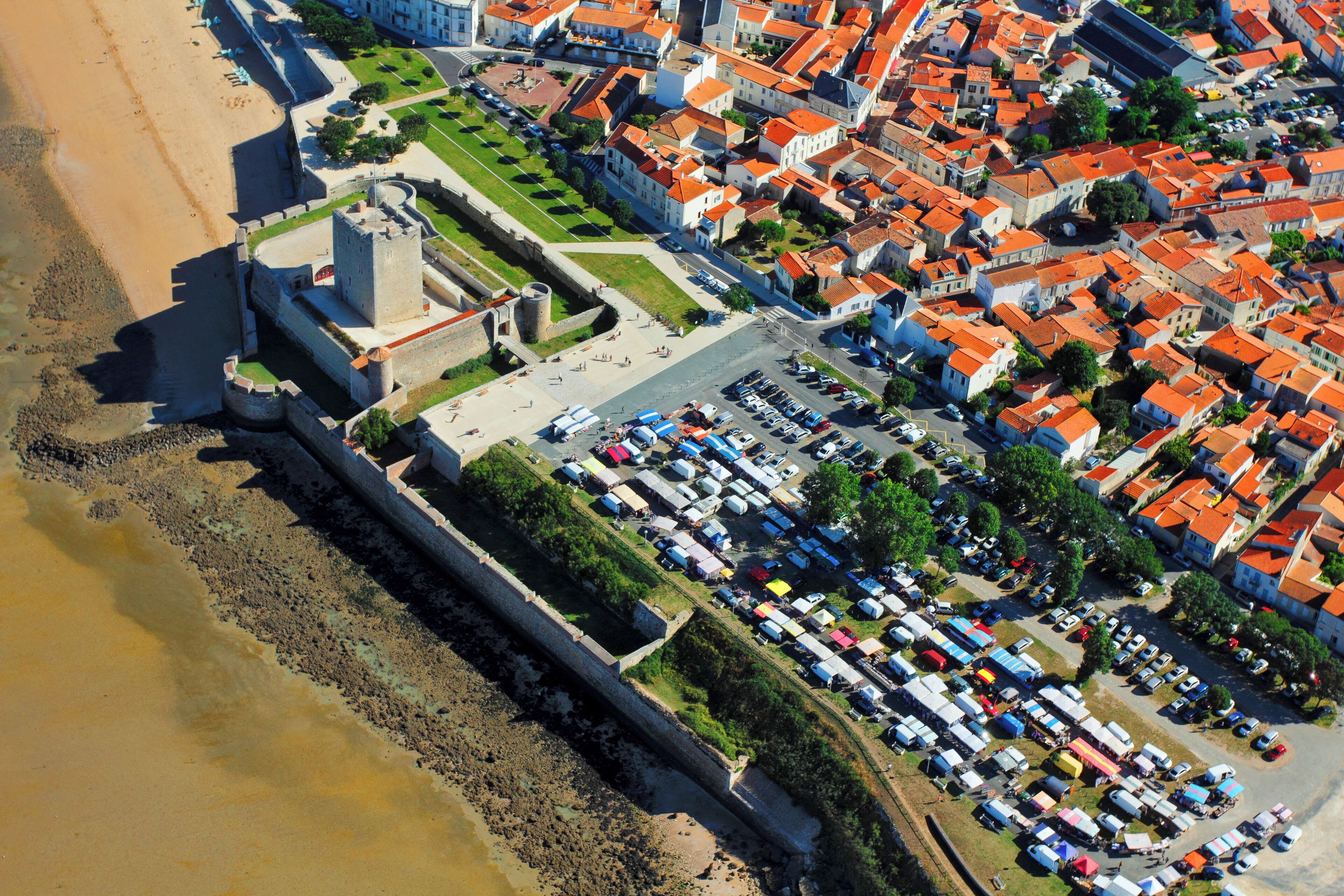 Vista aérea de un pueblo costero con una fortaleza histórica, un puerto deportivo y una zona densamente poblada con edificios de colores.