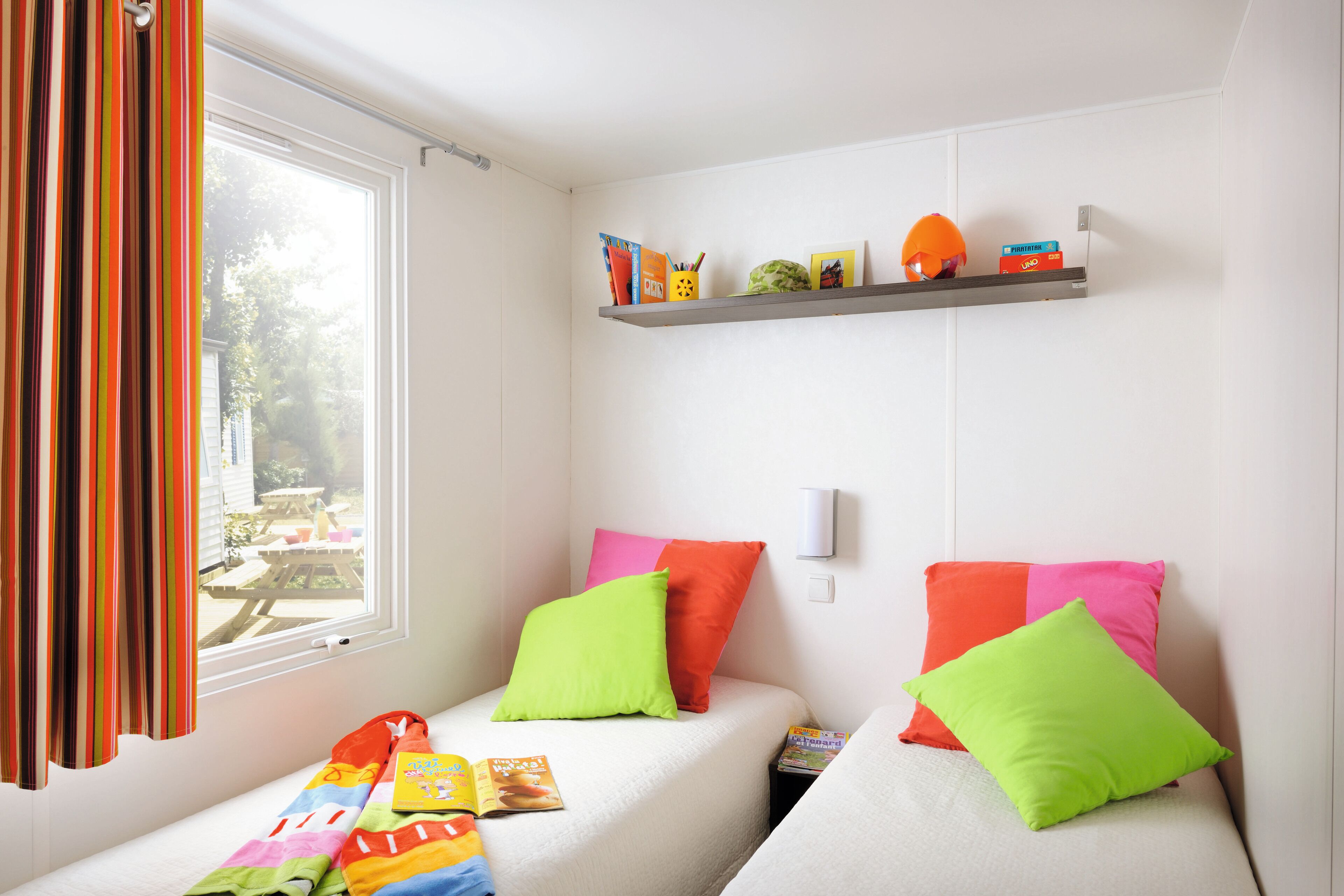 A small, bright room with two beds, colorful pillows, a shelf with books and a helmet, and a window with curtains.