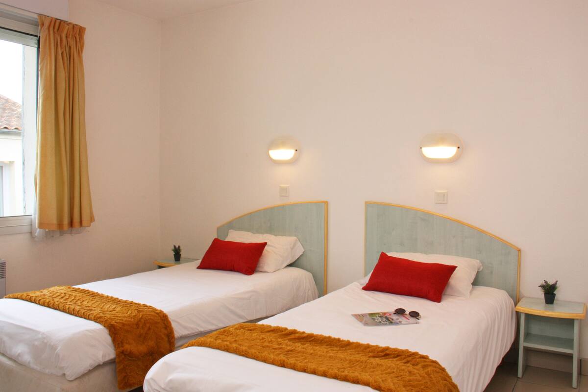 A hotel room with two single beds, each with a red pillow and a white blanket. There are two bedside tables with small potted plants.