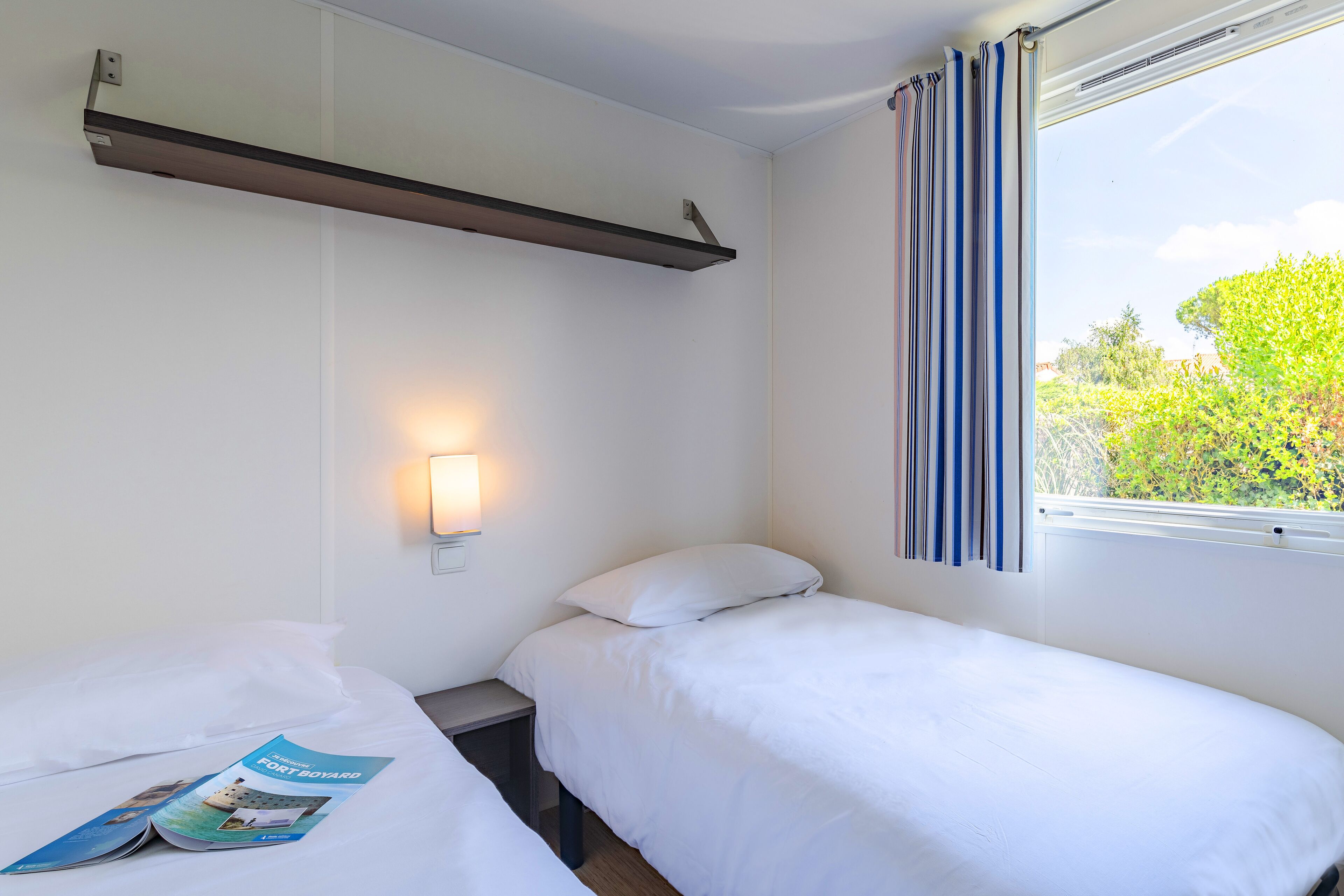Une chambre d’hôtel avec deux lits simples, une table de chevet, une étagère fixée au mur, une fenêtre avec des rideaux et une lampe fixée au mur.