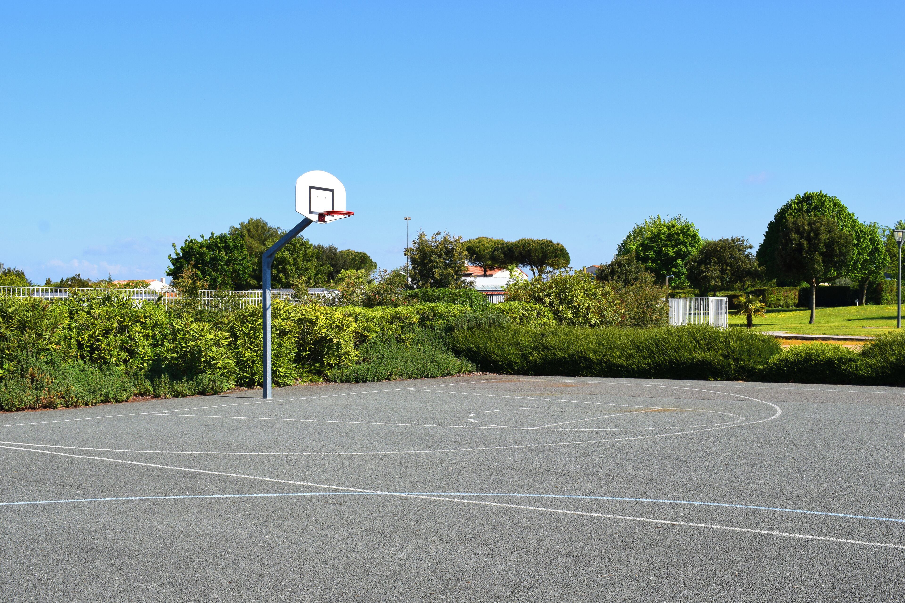 Un campo da basket all'aperto con canestro, circondato da vegetazione e un cielo sereno.