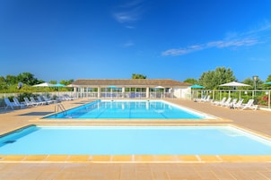 Une piscine rectangulaire, bordée d’une terrasse carrelée, entourée de chaises longues et de parasols, sous un ciel bleu dégagé.