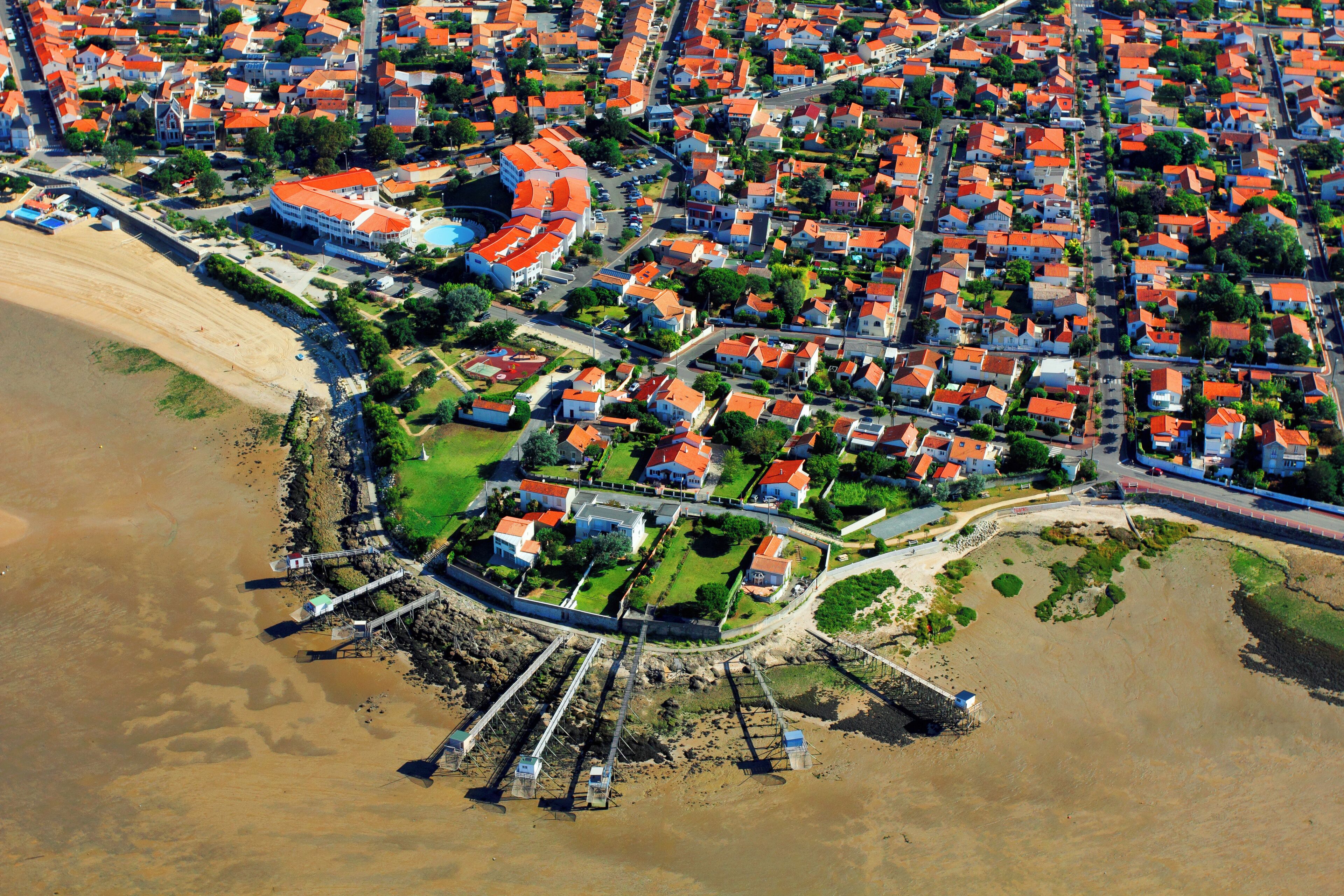 An aerial view of a coastal town with red-roofed houses, a sandy beach, and a pier.