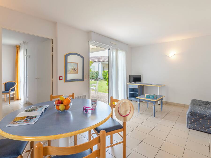 A dining area with a round table, chairs, and a bowl of fruit. There is a television and a small table with a TV stand in the background.