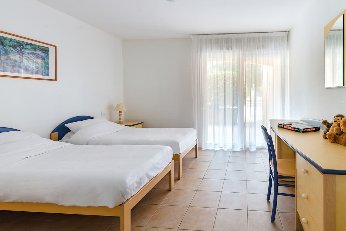 Ein Zimmer mit zwei Betten, einem Schreibtisch, einem Stuhl, einer Lampe und einem Vorhangfenster.
