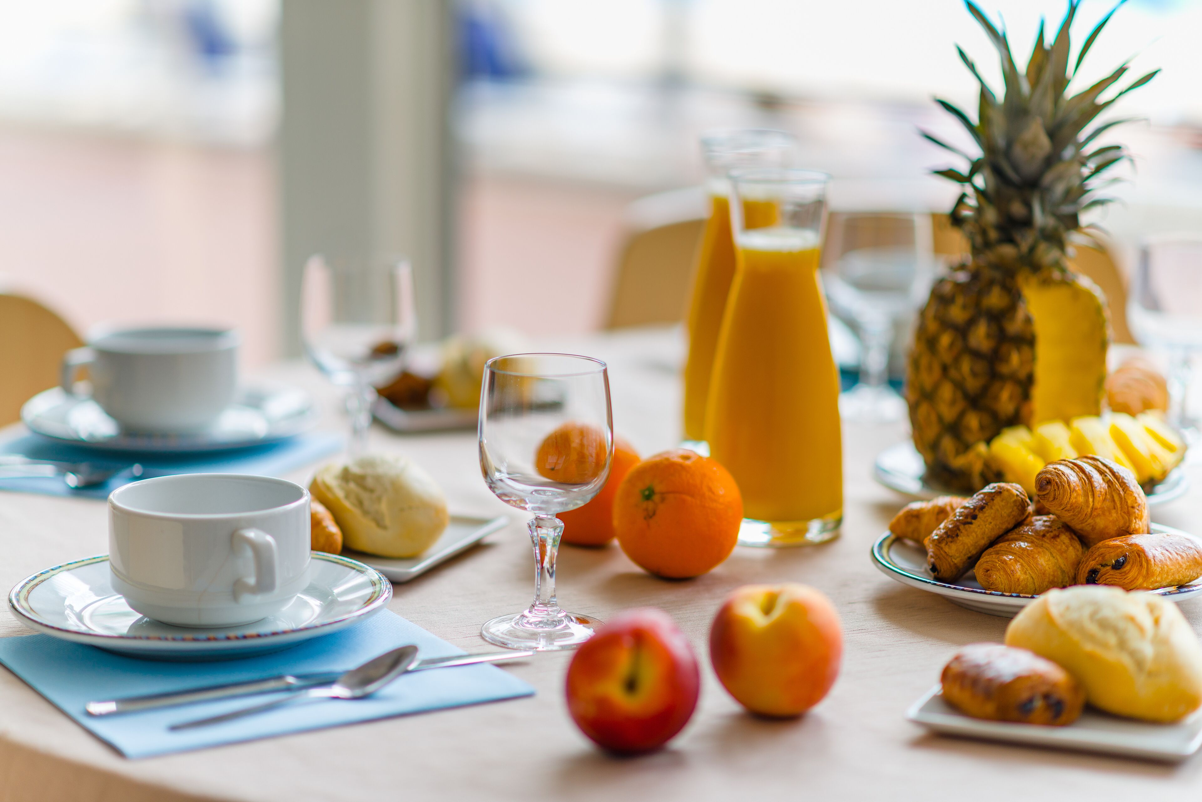 Una mesa de desayuno con una taza, un vaso, piña, naranjas y bollería.