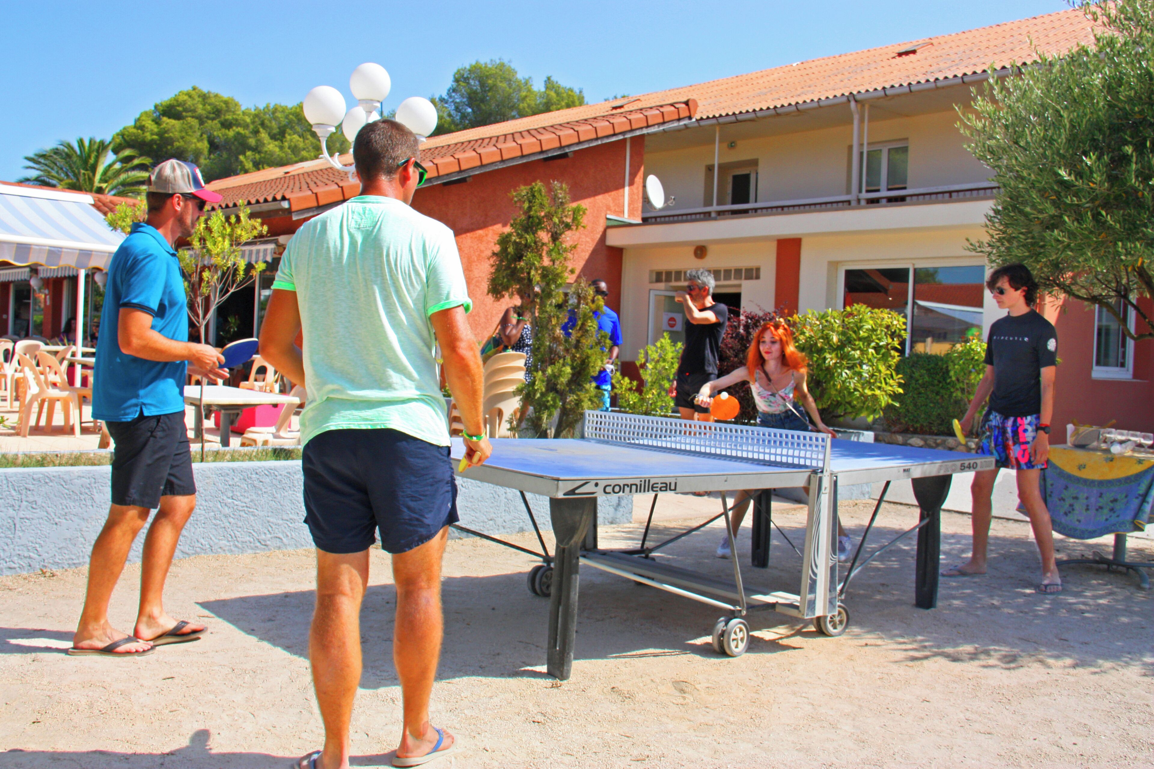 An outdoor table tennis setup with a metal table and net, surrounded by people playing and watching.