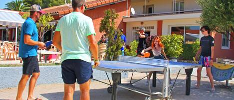 An outdoor table tennis setup with a metal table and net, surrounded by people playing and watching.