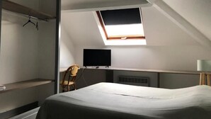 Una habitación pequeña con una cama, una silla, un televisor en la pared y un tragaluz.