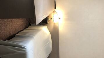 A room with a bed covered in white sheets, a wall-mounted light fixture, and a partial view of a carpeted floor.