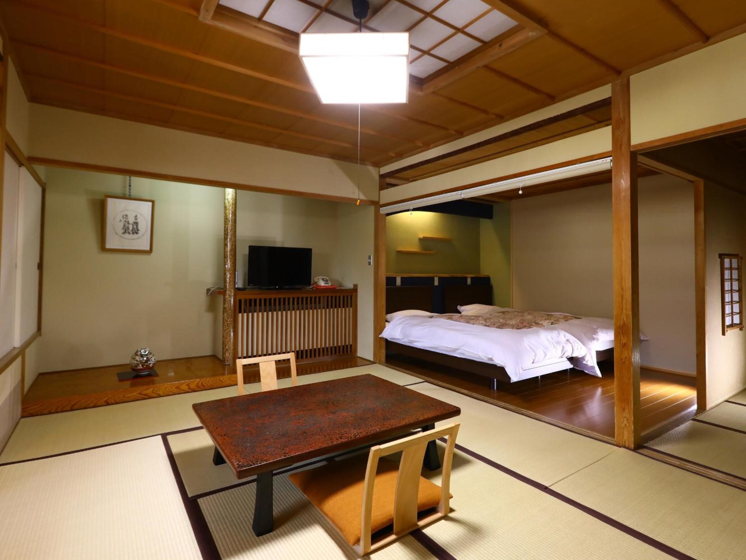A traditional Japanese room with tatami flooring, a low wooden table, a bed with a wooden headboard, and a television on a low stand.