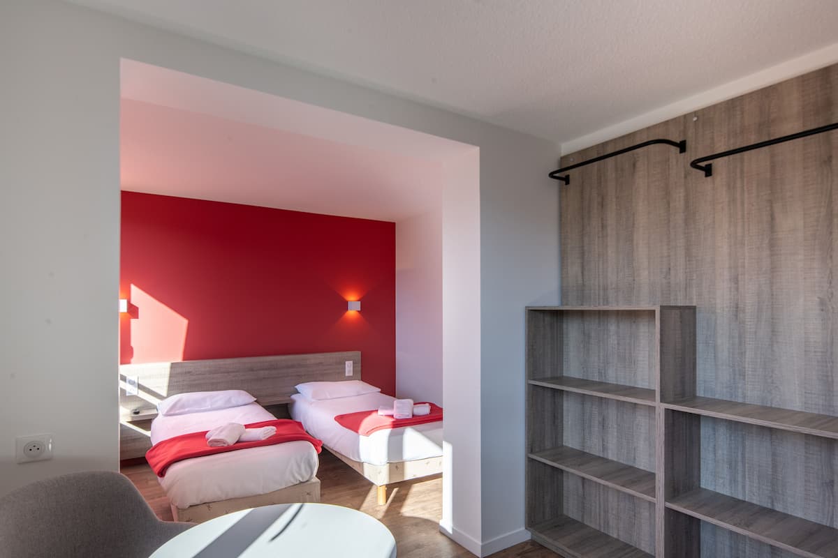 Camera d'albergo con due letti, un mobiletto a scaffali in legno e una parete decorata di rosso.