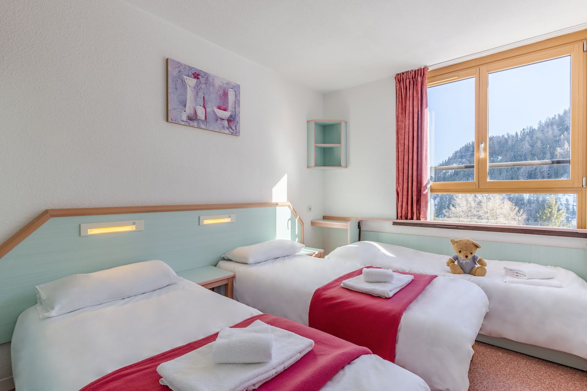 Camera d'albergo con due letti, una finestra vista alberi, un quadro al muro e un orsetto su un letto.