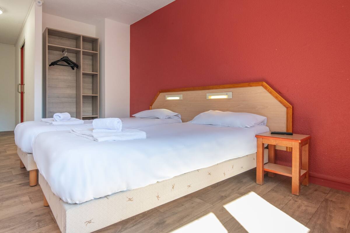 Un letto matrimoniale con lenzuola bianche e una testiera in legno, un comodino in legno con un telecomando, e un armadio a muro con spazio per appendere i vestiti.